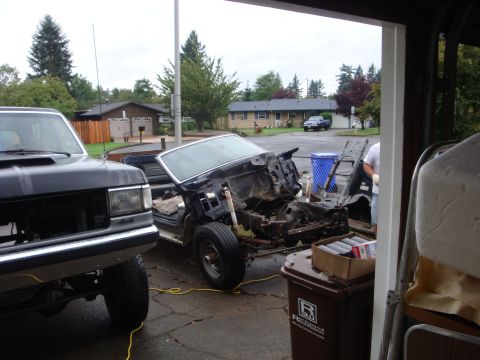 1989 Ford Bronco - A stripped Ford truck sits in a driveway mid-teardown with the front clip, fenders, hood and engine removed, exposing the bare frame rails,…
