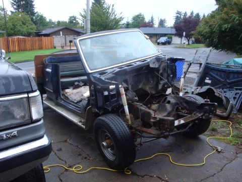 1989 Ford Bronco - A dark blue Ford truck partway through a frame-off teardown in a driveway, its cab and bed removed to expose the bare frame and front suspe…