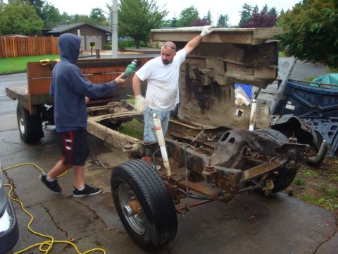 1989 Ford Bronco - Two people work on a bare full-size truck frame in a driveway, the cab tipped up on end to expose the dirty firewall and floor while the ro…