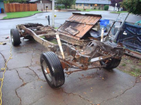 1989 Ford Bronco - A bare truck frame stripped to the rails sits on wheels in a wet residential driveway, with front coil-spring suspension, radius arms, whit…