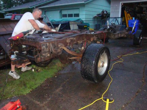 1989 Ford Bronco - Bare truck frame stripped to the rails sitting in a driveway with a man in a white shirt leaning over it, leaf springs and rear axle still…