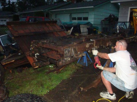 1989 Ford Bronco - A person in a white T-shirt and denim shorts crouches with a corded grinder, throwing sparks as they cut on a heavily rusted truck frame ra…