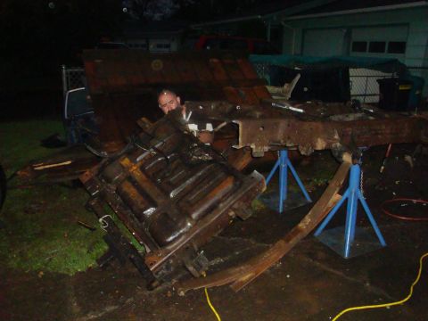 1989 Ford Bronco - Night shot of a truck teardown in a driveway: a rusty body tub and floor pan lie tipped on their side in the foreground with a leaf spring…