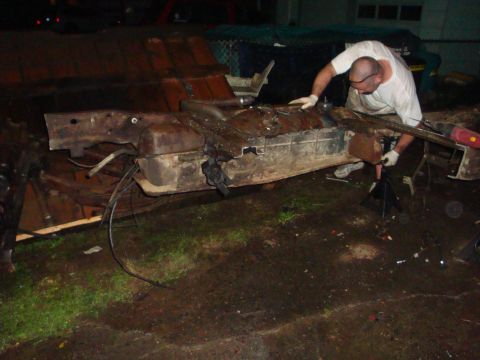 1989 Ford Bronco - Night shot of a man in a white t-shirt and work gloves prying at a rusty truck floor pan and fuel tank assembly propped on stands and wood…