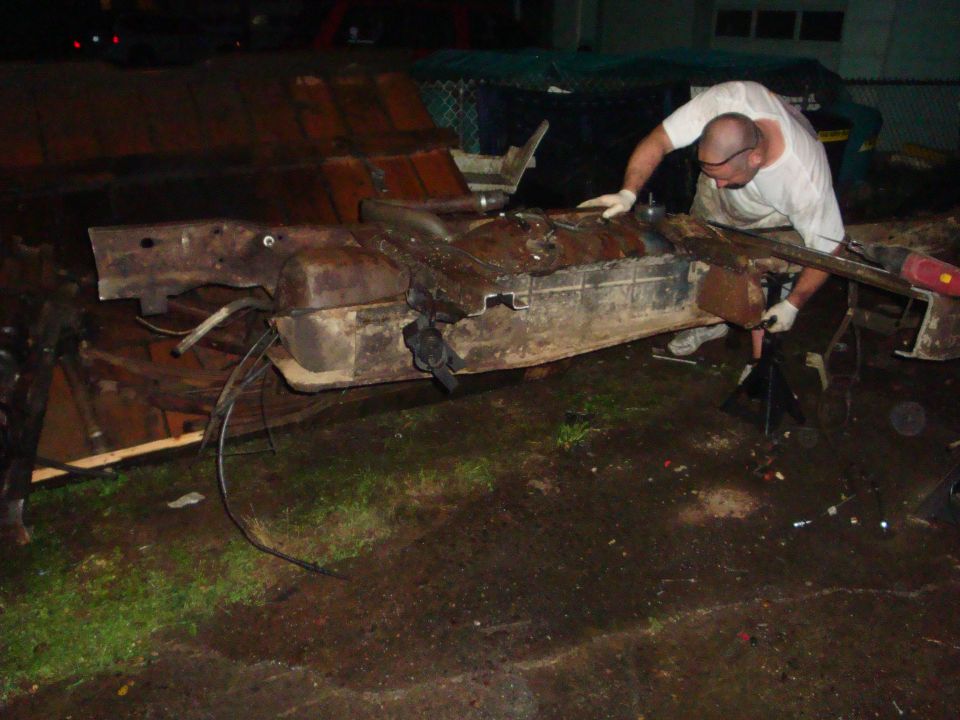 Man dismantling rusty truck floor pan and fuel tank on stands outdoors at night