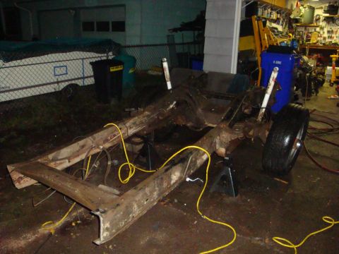 1989 Ford Bronco - A bare, surface-rusted truck frame sits on jack stands outside an open garage at night, stripped of body and drivetrain with only the rear…