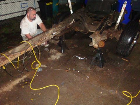 1989 Ford Bronco - A stripped rusty truck frame sits on jack stands in a driveway with the body removed, exposing the rear leaf springs, axle, white-bodied sh…