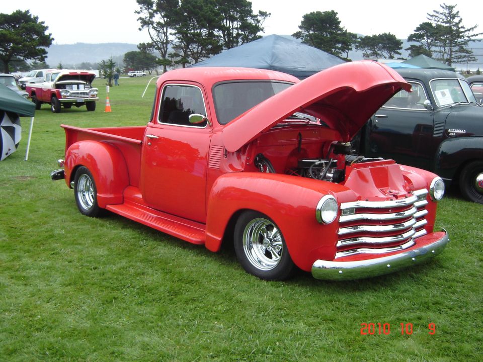 Red pickup truck with hood open, chrome grille, and custom wheels. Another dark pickup is parked behind it