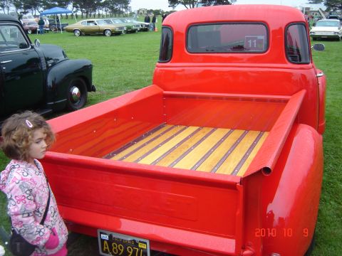 1993 Ford Bronco - A young girl stands next to the polished wooden bed of a bright red pickup truck at an outdoor car show.