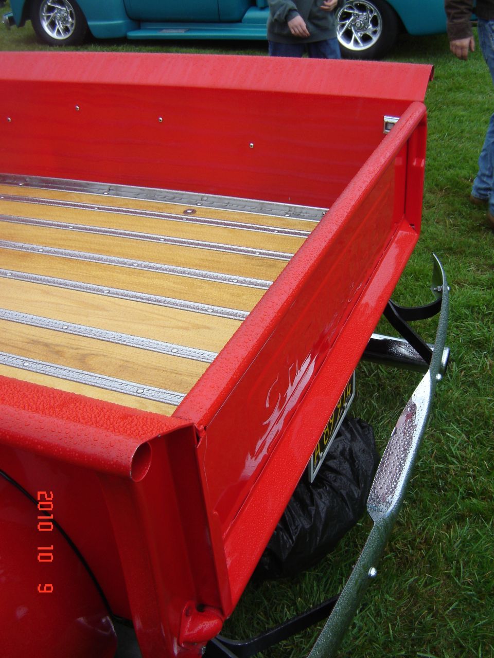 Red pickup truck bed with wooden slats and chrome bumper. Teal vehicle in background