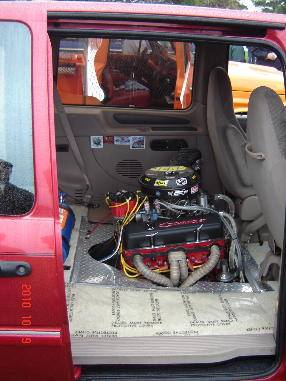 Engine bay of a red vehicle with a Chevrolet V8 engine installed. Another vehicle is visible outside