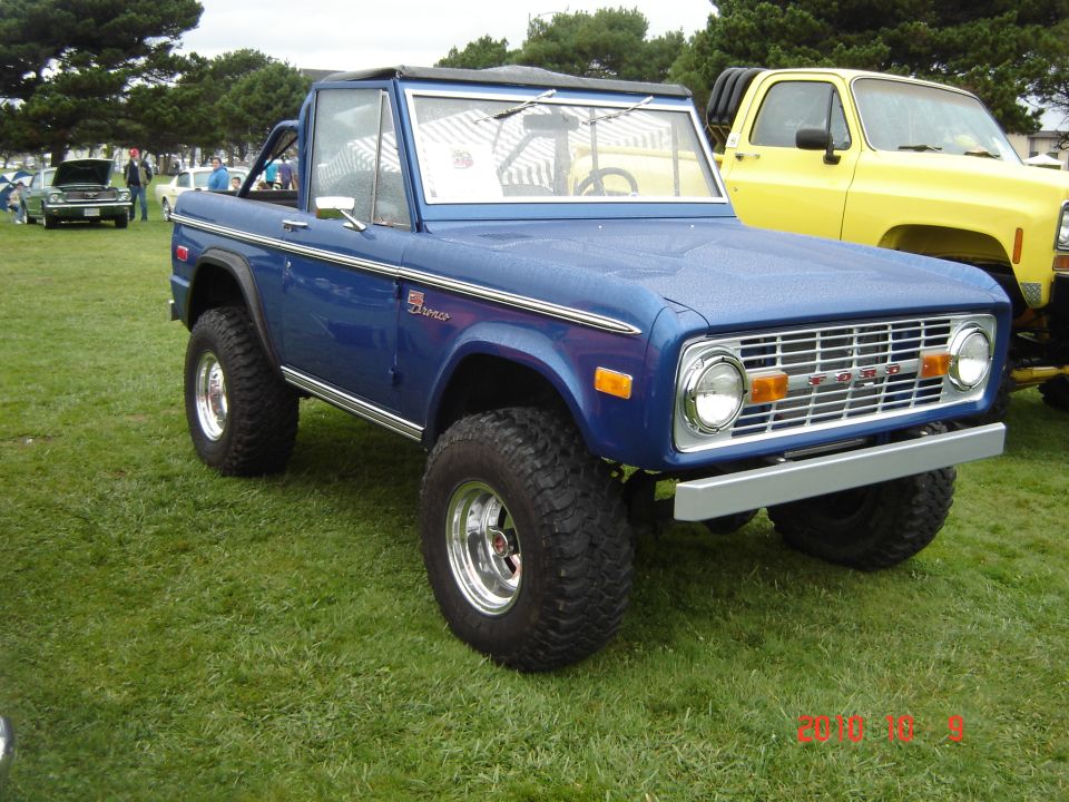 Blue Ford Bronco with aftermarket wheels and tires parked on grass next to a yellow truck