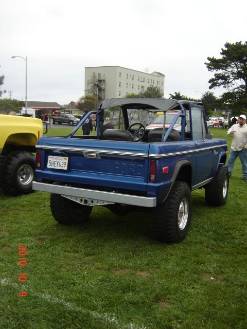 1993 Ford Bronco - A blue Ford Bronco is parked on grass, showing its rear end and lifted suspension, with another vehicle to its left.
