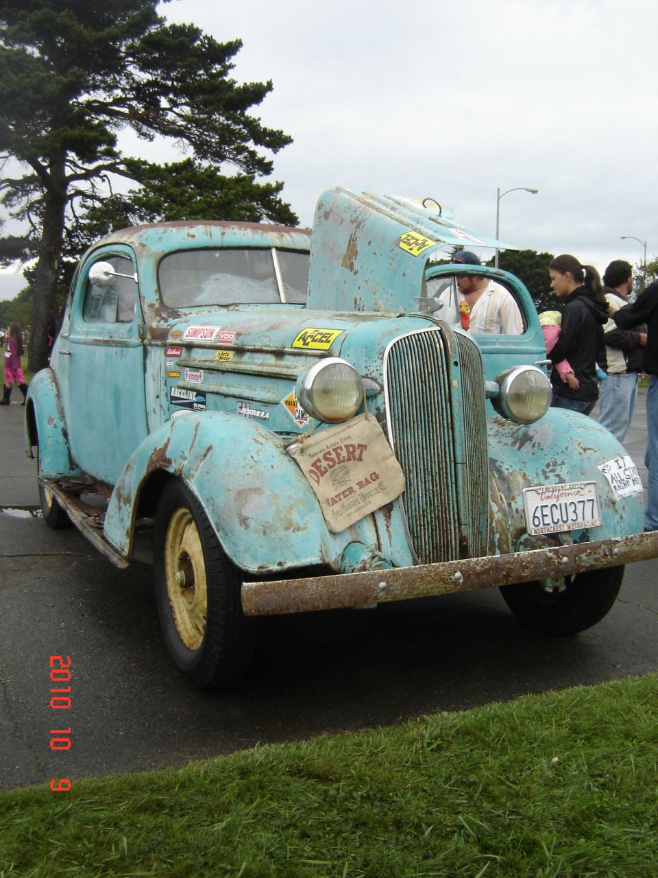 Distressed light blue vintage truck with hood up, showing a fabric bag labeled 'Desert Water Bag'