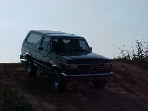 1989 Ford Bronco - A black Ford Bronco navigates an off-road trail under a hazy sky.