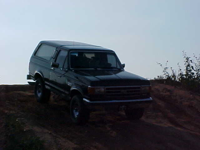 Black Ford Bronco driving up a dirt hill. The grille and headlights indicate it's from the late 1980s