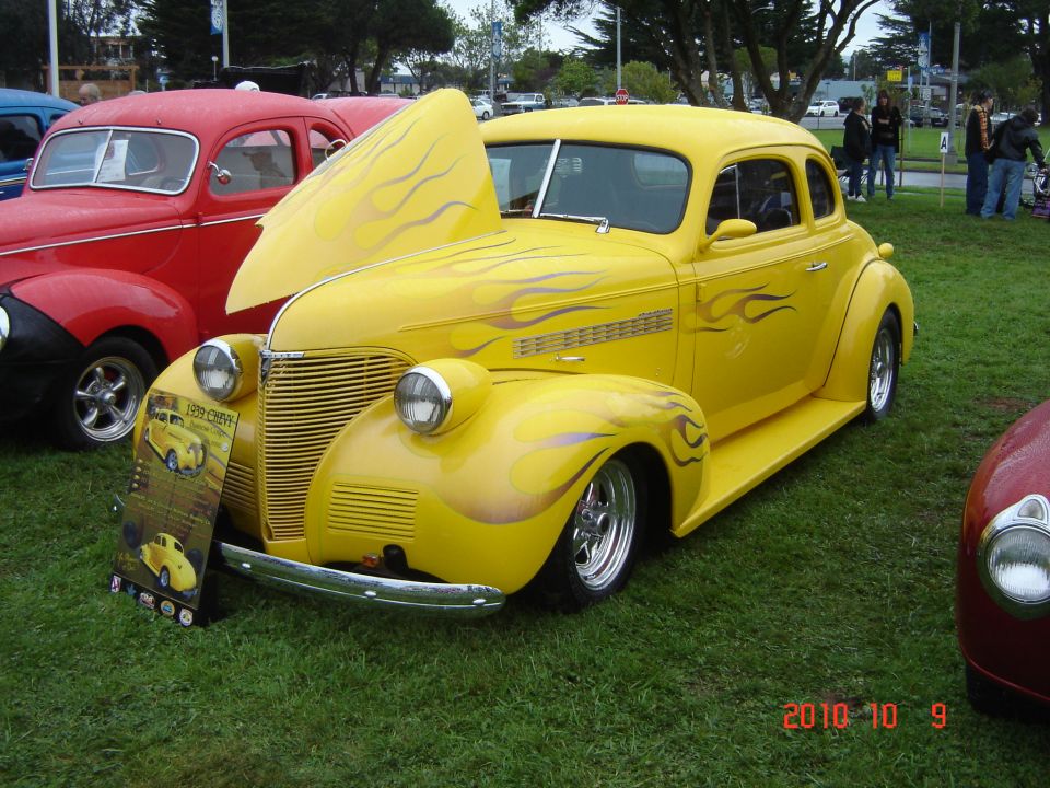 Yellow hot rod with flame graphics and hood open, parked on grass next to a red car. A sign in front of it says '1939 CHEVY'