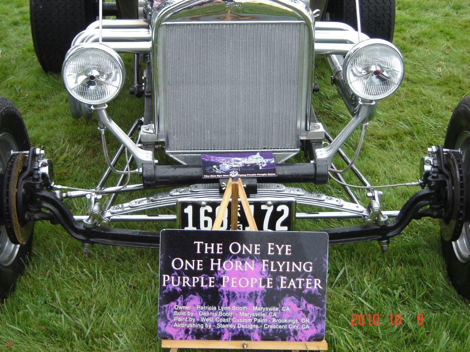 Front view of a custom hot rod with a chrome grille, dual headlights, and a license plate reading 16A 72. A sign in front names it 'The One Eye One Horn Flying Purple People Eater'