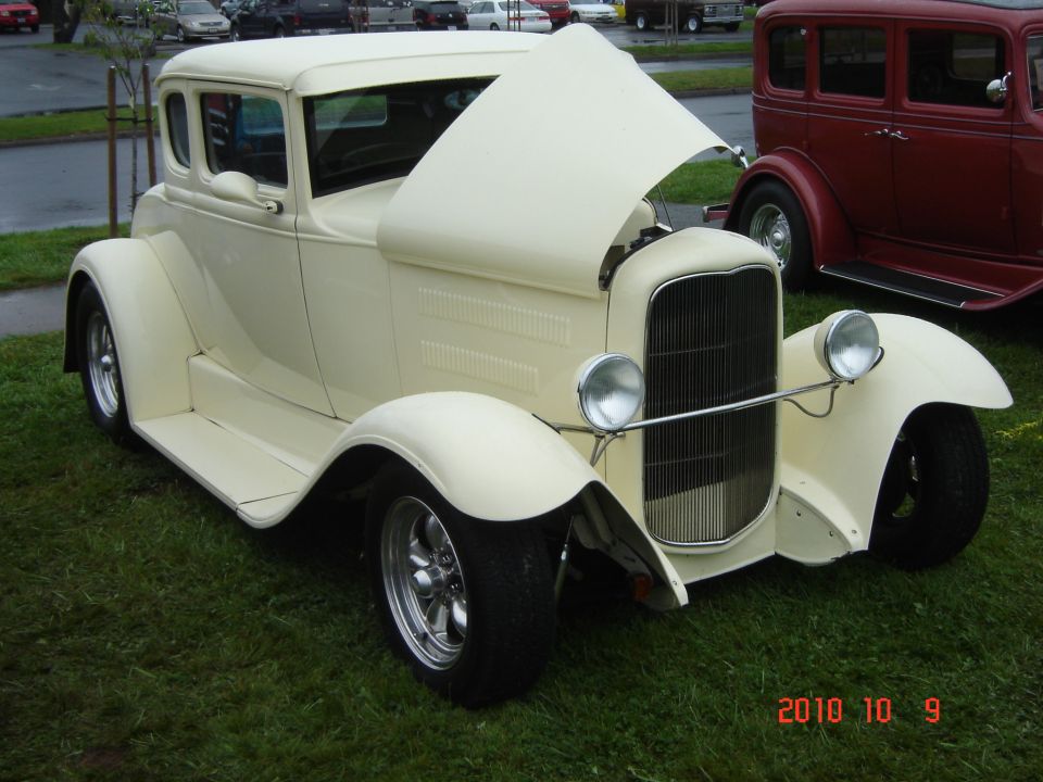 Cream-colored 1930s hot rod coupe with hood open, parked on grass next to a red sedan