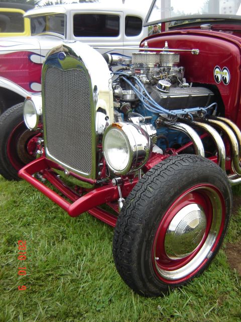 1993 Ford Bronco - A red hot rod with a chrome grille and exposed engine is parked on grass next to another car.