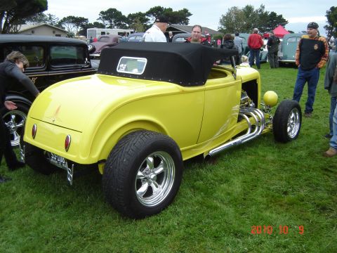 1993 Ford Bronco - A bright yellow hot rod roadster is displayed on a grassy field with other vehicles and people in the background.
