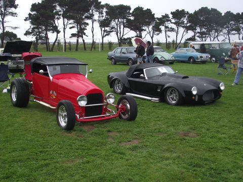 1993 Ford Bronco - Two classic cars, a red hot rod and a black sports car, are displayed on a grassy field with other vehicles present.