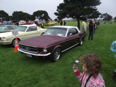 1993 Ford Bronco - A young girl photographs a maroon convertible parked on a grassy field during an outdoor car show.