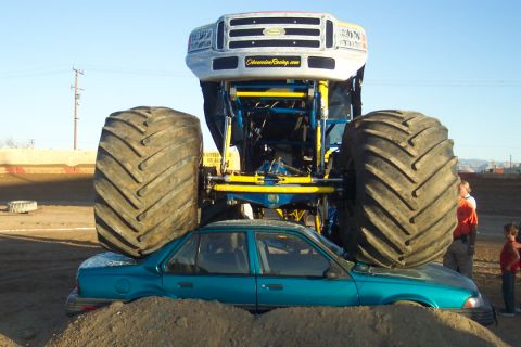 1995 Ford Bronco - A monster truck with large tires is shown crushing a teal sedan in a dirt arena.