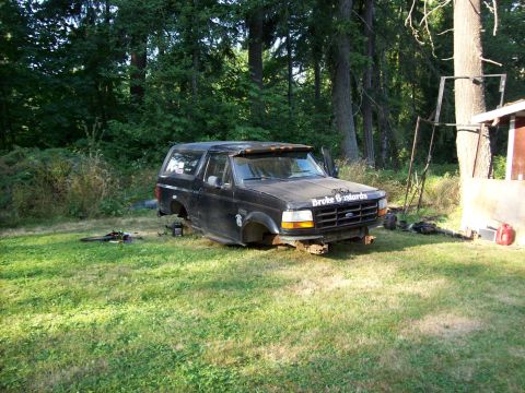 1995 Ford Bronco - A black Ford Bronco sits on its frame in a grassy yard, stripped of its wheels and surrounded by trees.