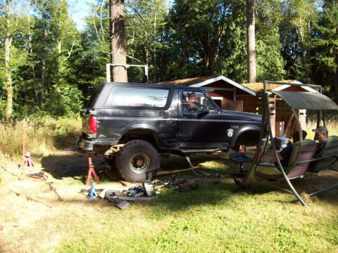 1995 Ford Bronco - A black Ford Bronco is lifted on jack stands in a yard, with tools scattered around and a swing set nearby.