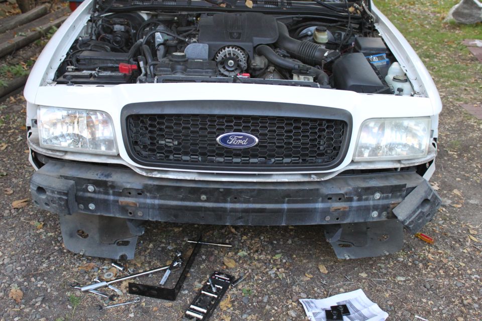Front of a white Ford Ranger with grille removed and bumper hardware on ground