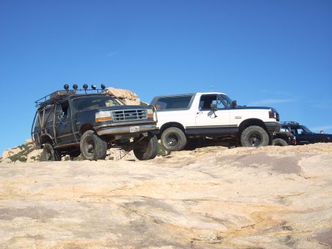 1989 Ford Bronco - Two Ford Broncos, one modified with a roll cage, are parked on rocky terrain under a clear blue sky.