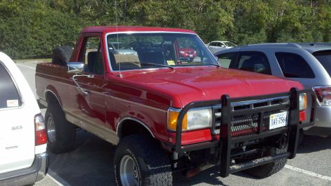 1988 Ford Bronco - A red pickup truck is parked next to other vehicles in a parking lot, a prominent brush guard on the front.