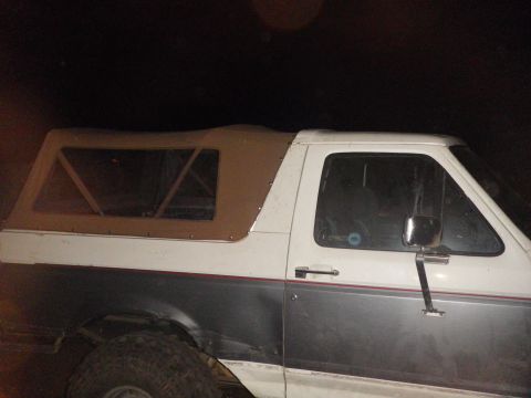 1991 Ford Bronco - Night shot of the driver's side of a two-tone white-over-gray Ford Bronco fitted with a tan aftermarket soft top over the rear tub, showing…