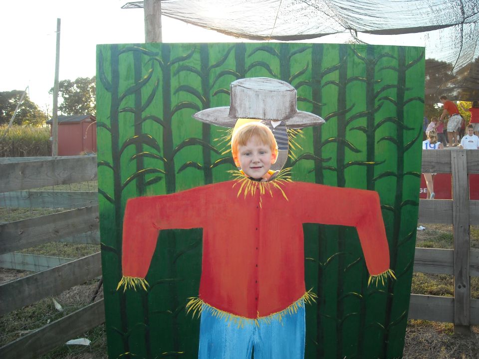 kid's head sticking out of a scarecrow cutout with corn stalks painted behind