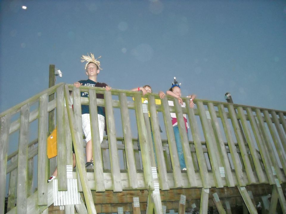 Three children wearing party hats stand on a wooden deck against a dark sky
