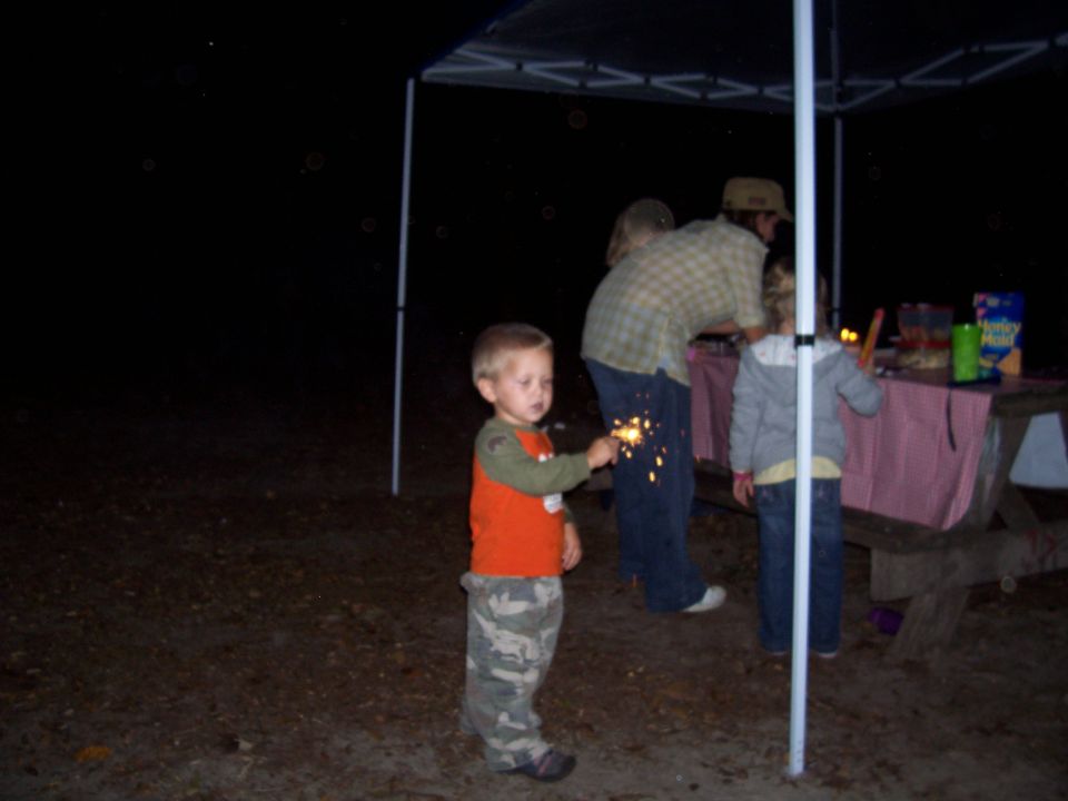 Kid holding a sparkler, people at picnic table under canopy at night