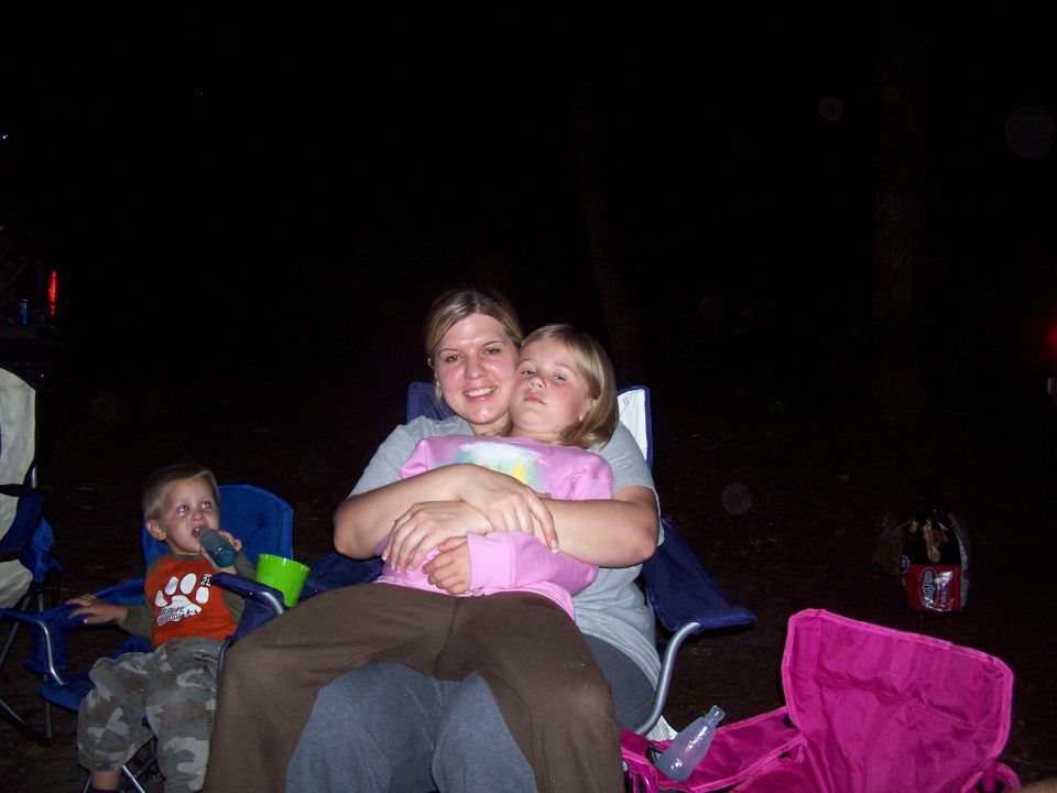 Family sits in camping chairs at night, with a woman holding two children. A pink cooler is in the foreground