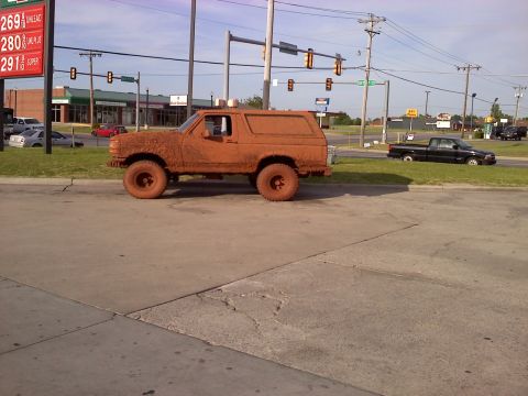 1994 Ford Bronco - A heavily mud-covered SUV is parked at a gas station, obscuring its exact make and model year.