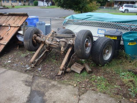 1989 Ford Bronco - A bare truck frame with axles, leaf springs and steering linkage still attached sits on its wheels in a grassy strip beside a chain-link fe…