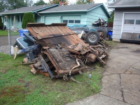 1989 Ford Bronco - A pile of stripped truck parts heaped on a front lawn, including a rusty frame rail, fuel tank, wiring, weathered wood bed planks, and a re…