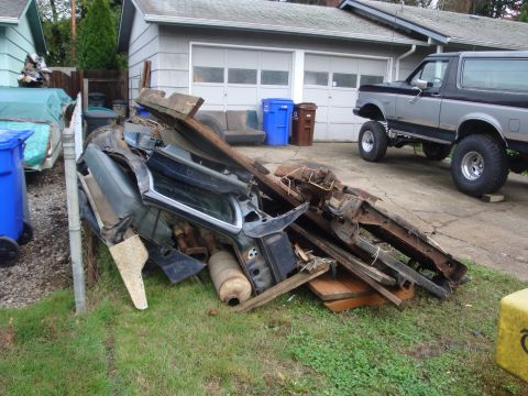 1989 Ford Bronco - A scrap pile of removed truck parts heaped on a lawn beside a driveway, a teal cab shell section, muffler, bed panels, rusty frame rails an…
