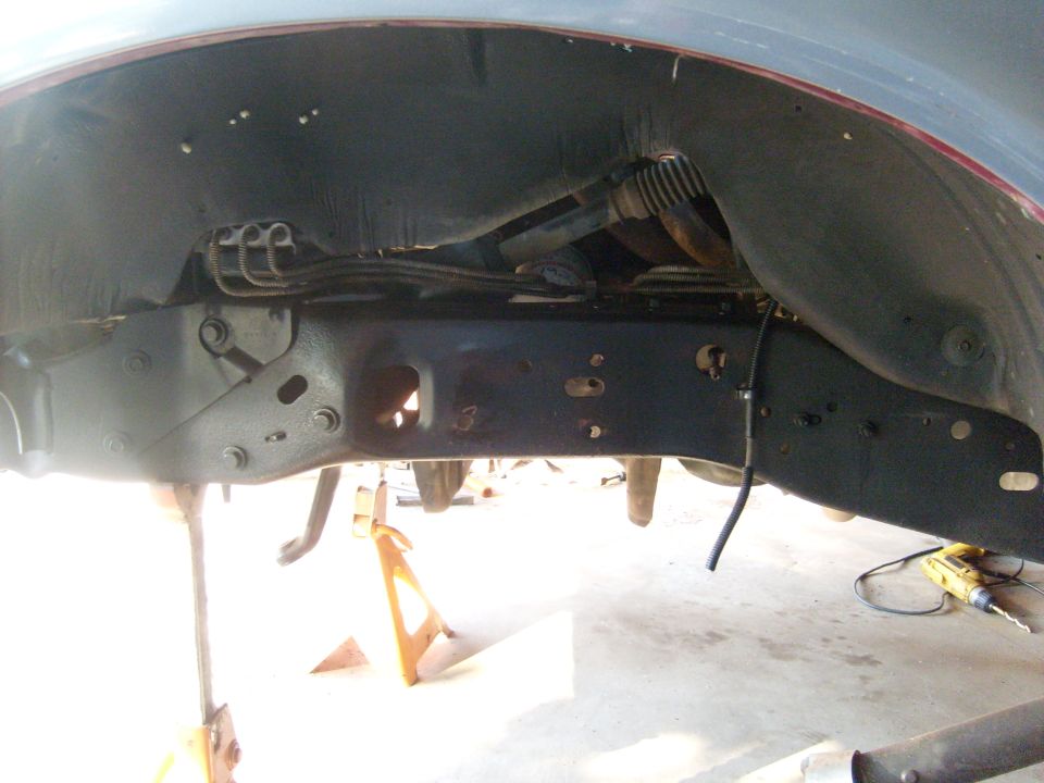 Underside of a vehicle on jack stands, showing the frame and suspension components. A drill is visible on the right