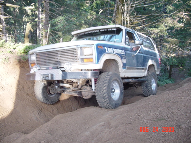 Blue and white lifted Ford Bronco articulating over a dirt bank on a wooded trail