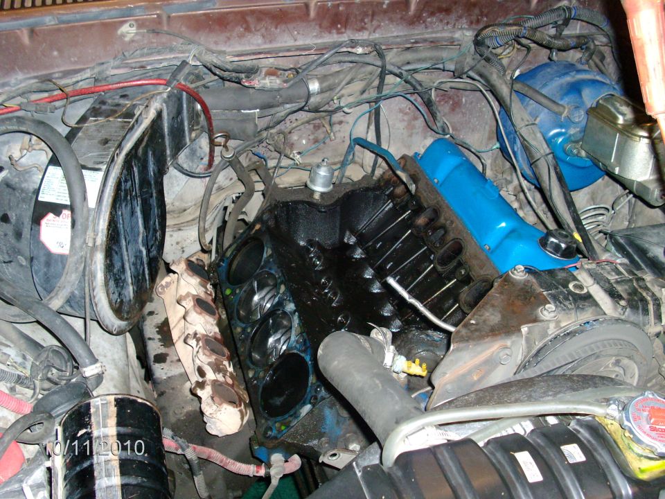 Engine bay teardown with cylinder head removed exposing bores, blue valve cover on opposite bank