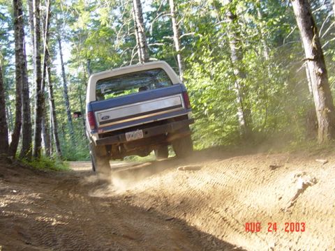 1989 Ford Bronco - 1989 Ford Bronco navigates a dirt path through a wooded area, kicking up dust.