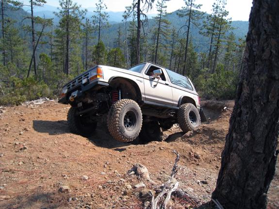 Lifted tan Ford Bronco II articulating out of a dirt ditch on a forested ridge trail