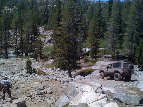 1990 Ford Bronco II - Wide trail scene of a lifted tan Bronco II picking its way across broken granite slabs in a Sierra pine forest, viewed from the driver's si…
