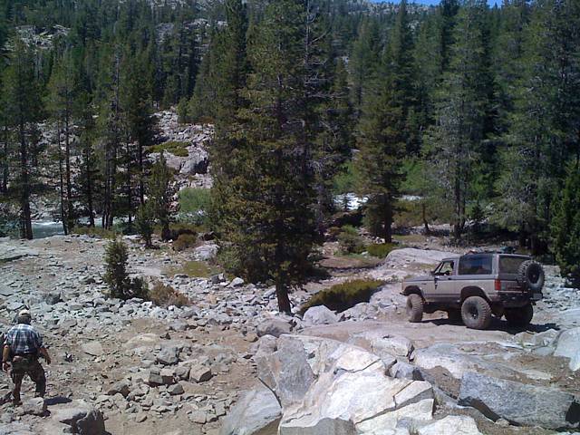 Lifted tan Bronco II on granite rocks in a pine forest with a spotter walking ahead