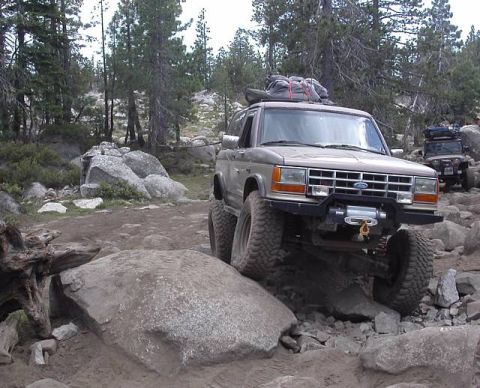 1990 Ford Bronco II - Lifted tan two-tone Ford Bronco II crawling over granite rocks on a high-country pine trail, seen from the front-right three-quarter with a…